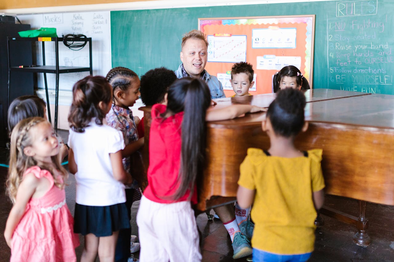 The Art of Drawing Readers In: Your attractive post title goes here Teacher guiding children in music class around a piano, fostering creativity and diversity.